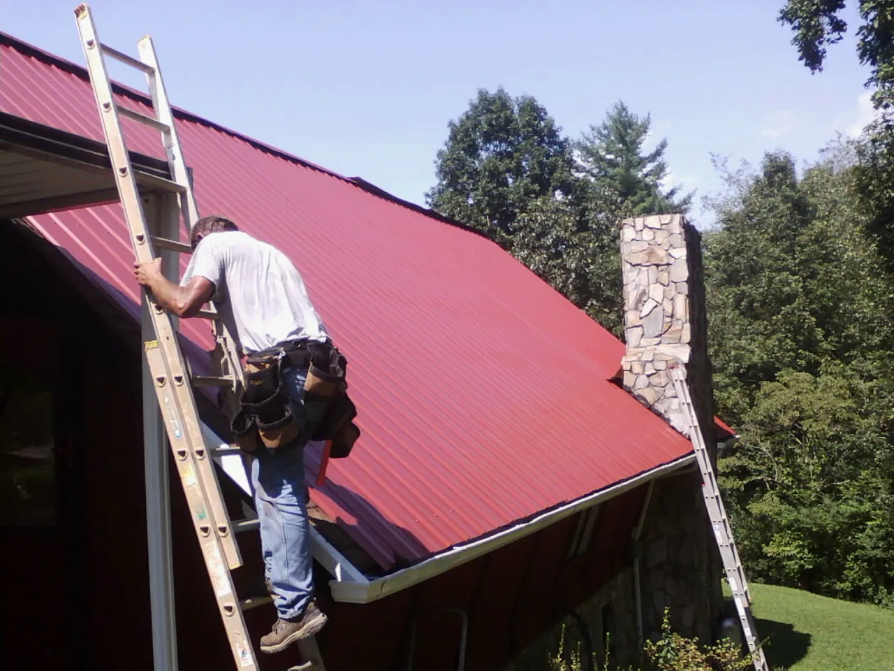 Roofer on a ladder beside a red metal roof and stone chimney — Soffit & Fascia Repair by Roofing Brothers in Feasterville Trevose